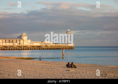 A couple sit alone on the beach enjoying the calm sea and evening sunlight with the pier in the background at Bournemouth, UK Stock Photo