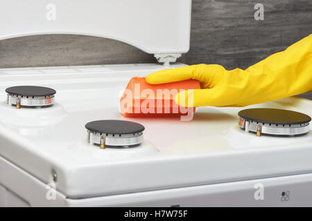Woman's hand in yellow rubber protective glove cleaning white gas stove with orange sponge on gray background Stock Photo