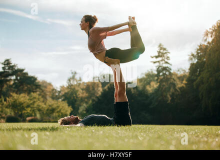 Fit young couple doing acro yoga. Man lying on grass and balancing woman in his feet. Acrobatic balance in park. Stock Photo