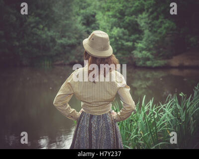 A young woman wearing a safari hat is standing by a pond in the forest on a sunny summer day Stock Photo