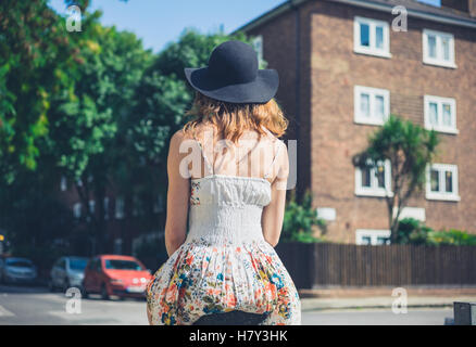 A young woman wearing a dress and a hat is sitting on a bollard in the street on a sunny day Stock Photo