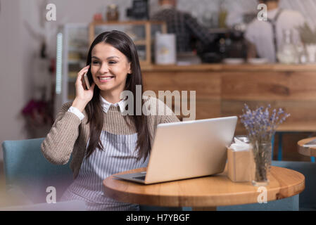 Smiling cute woman using laptop and talking on cellphone. Stock Photo