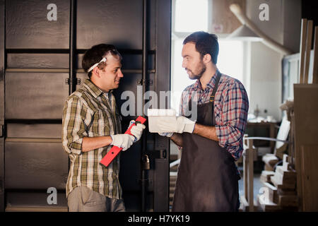 Two carpenters are talking while holding papers and ruler. One of them is wearing safety mask. Carpentry shop background Stock Photo