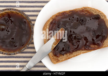 Top close view of plum preserves spread on a piece of wheat bread atop a white plate with a butter knife Stock Photo