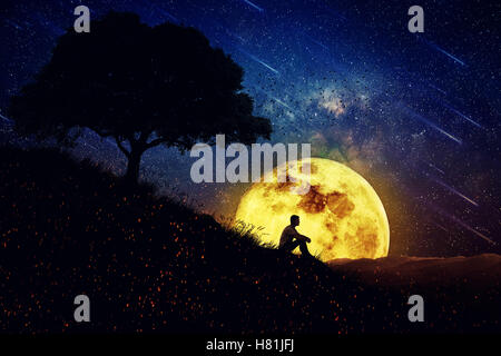Boy sit alone on a hill in the center of nature, over a full moon night background. Standing away from the crowd, waiting for th Stock Photo
