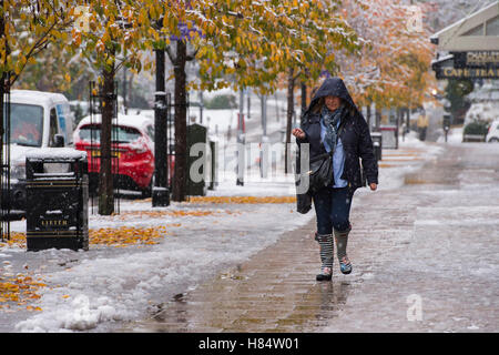 Ilkley, West Yorkshire, UK. 9th November 2016. It is snowing and a lady (wearing boots and a winter coat with hood up) is walking past shops on The Grove - Ilkley's first snowfall of 2016. Credit:  Ian Lamond/Alamy Live News Stock Photo