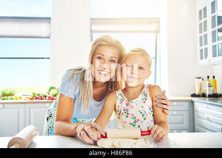 Smiling happy young mother and daughter learning to bake posing arm in arm at the kitchen counter Stock Photo