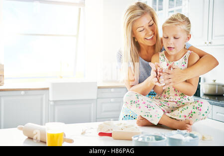 Little girl seated cross legged on kitchen table getting help to mash bread dough with enthusiastic woman Stock Photo