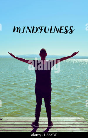 a young caucasian man seen from behind with his arms in the air saluting the new day in front of the sea and the text mindfulnes Stock Photo