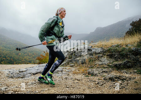 runner middle-aged woman running in rain on a mountain trail with walking poles during Crimea mountain marathon Stock Photo