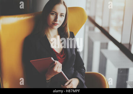 Portrait of young serious successful woman entrepreneur in a red dress and jacket sitting on yellow armchair with digital tablet Stock Photo
