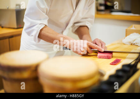 A chef working in a small commercial kitchen, an itamae or master chef slicing fish with a large knife for making sushi Stock Photo