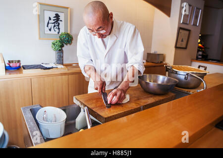 A chef working in a small commercial kitchen, an itamae or master chef slicing fish with a large knife for making sushi Stock Photo