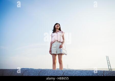 Young woman in shorts and pink shirt standing alone in open space, with a large suspension bridge in the background Stock Photo