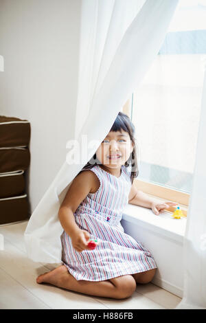 Family home. A girl playing by a window, hiding behind the net curtain. Stock Photo