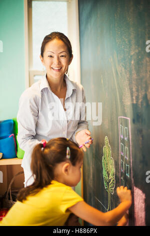 A child writing on the chalkboard and a teacher beside her. Stock Photo
