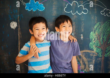 Children in school. Two boys standing with their arms around each other's shoulders by a chalkboard. Having fun at school. Stock Photo