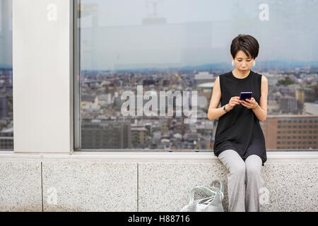 A woman seated by a window with a view over a large city with her back to the view, using her smart phone. Stock Photo