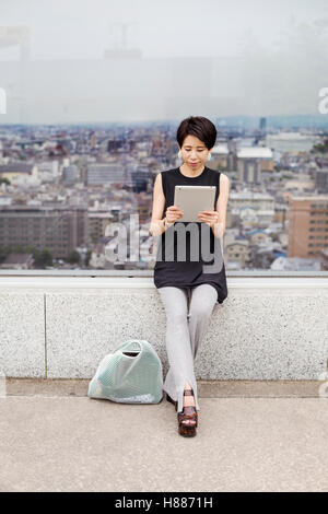 Woman using a digital tablet, with her back to the view over a large city. Stock Photo