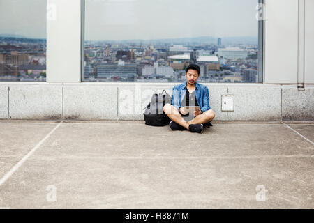 A man sitting on the floor using his smart phone, in front of a viewing point window on to a city. Stock Photo