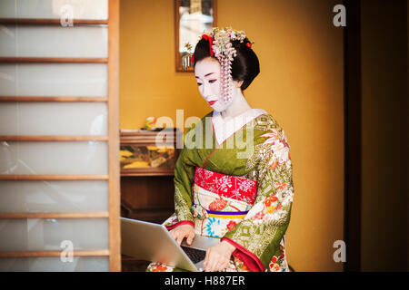 A woman dressed in the traditional geisha style, wearing a kimono and obi, with an elaborate hairstyle and floral hair clips, with white face makeup with bright red lips and dark eyes using a laptop computer. Stock Photo