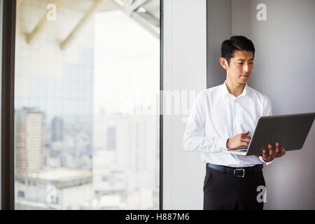 A business man standing by a window with a city view, using his laptop. Stock Photo