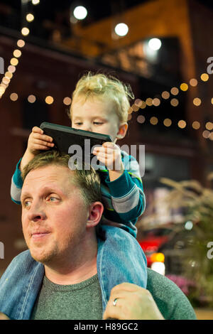 Detroit, Michigan - Two-year-old Adam Hjermstad Jr. looks at a cell phone while riding on the shoulders of his dad. Stock Photo