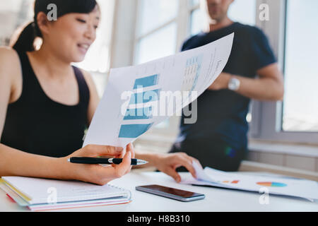 Woman doing financial analysis at office with male colleague standing in background. Asian female holding documents while sittin Stock Photo