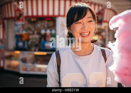 Shot of young asian woman at amusement park with cotton candy floss. Smiling female with candyfloss at fairground. Stock Photo