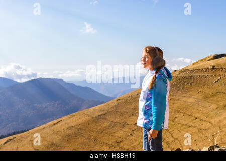 Girl with a pigtail, dressed in sports jacket and warm fluffy headphones, admires of autumn mountain landscape, Sochi, Russia Stock Photo