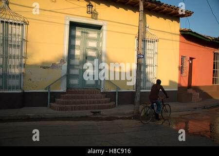 A Cuban man rides his bicycle past some brightly painted houses as the sun goes down in Trinidad Cuba Stock Photo