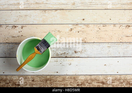 loaded paintbrush placed across a white paint kettle filled with green paint on  light multicolored floor boarding Stock Photo