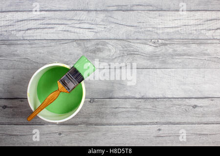 Loaded paintbrush placed across a white paint kettle filled with green paint on a grey shabby style wood floor Stock Photo