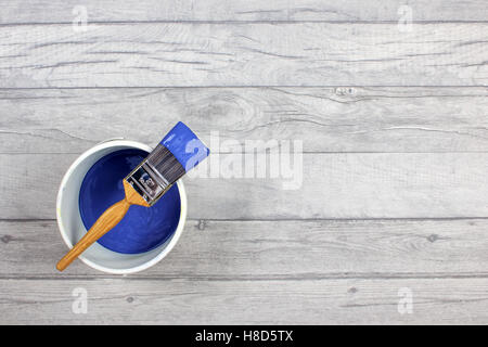 loaded paintbrush placed across a white paint kettle filled with dark blue paint on a grey shabby style wood floor Stock Photo