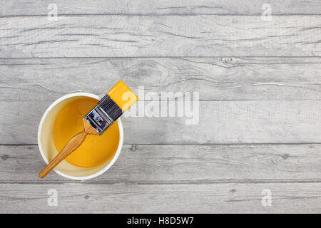 Loaded paintbrush placed across a white paint kettle filled with yellow paint on a grey shabby style wood floor Stock Photo