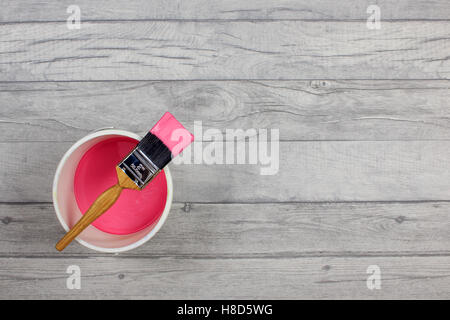 Loaded paintbrush placed across a white paint kettle filled with pink paint on a grey shabby style wood floor Stock Photo