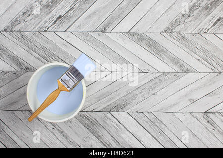 Loaded paintbrush placed across a white paint kettle filled with dark ligh blue paint on a grey and white herringbone stylefloor Stock Photo