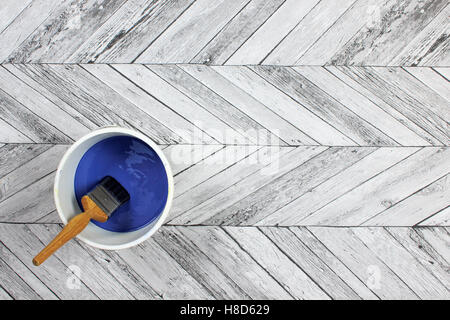 Paintbrush sitting in a white paint kettle filled with dark blue paint on a grey and white herringbone style wood floor Stock Photo
