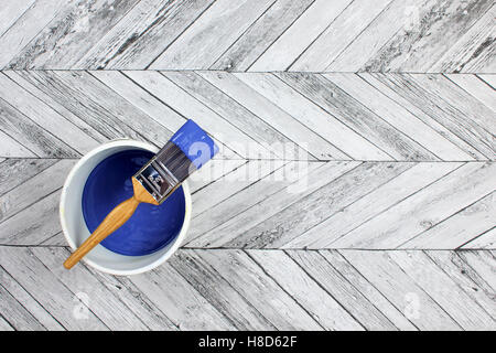 Loaded paintbrush placed across a white paint kettle filled with dark blue paint on a grey and white herringbone stylefloor Stock Photo