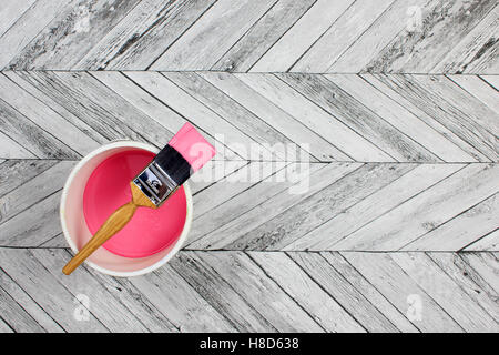 Loaded paintbrush placed across a white paint kettle filled with pink paint on a Grey and white herringbone wood floor Stock Photo