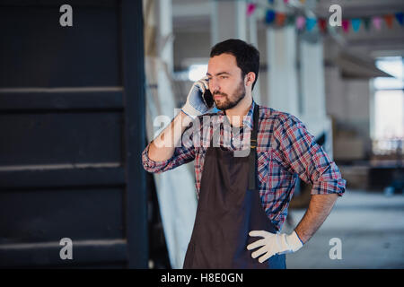 Smiling bearded man wearing gloves, owner of a small woodwork business, standing in his workshop studio, talking on  mobile phone Stock Photo