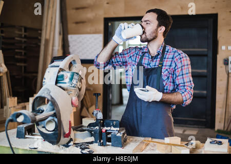 Coffee mug for good work. Thoughtful young male carpenter holding cup while standing near the wooden table with diverse working tools laying on it Stock Photo