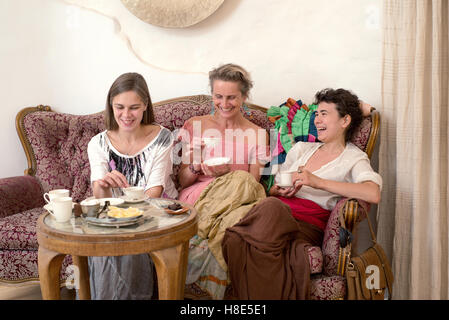 Three women in ethnic dresses sitting on an old couch and drinking tea Stock Photo