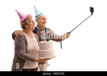 Happy seniors with party hats and a birthday cake taking a selfie with a stick isolated on white background Stock Photo