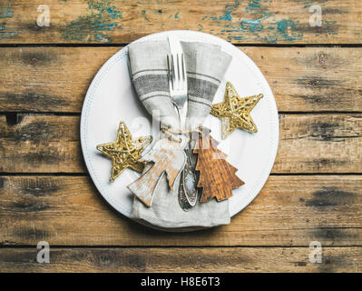 Christmas or New Year holiday rustic table setting. White plate with linen napkin, napkin holders, silverware and golden stars o Stock Photo