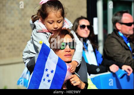 Little girl holding the flag of Honduras sits on her mother's shoulders while waiting to cheer on a runner on Michigan Avenue. Chicago, Illinois, USA. Stock Photo
