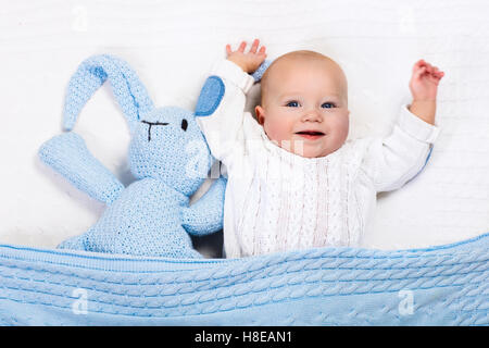 Funny little baby wearing a warm knitted jacket playing with toy bunny relaxing on white cable knit blanket in sunny nursery. Stock Photo