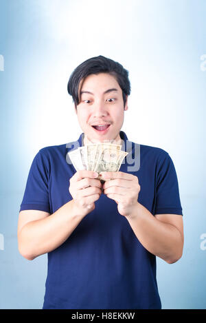 Successful Asian man. Happy young man holding money while standing and thumbs up, on blue background Stock Photo