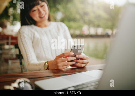 Shot of asian female using cellphone at outdoor cafe and smiling. Young female sitting at table with laptop reading text message Stock Photo