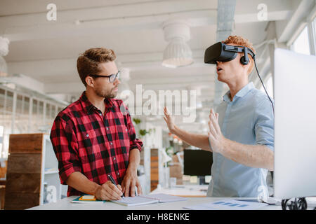 Shot of two young men testing virtual reality headset. Businessman wearing VR goggles and colleague writing notes in office. Stock Photo